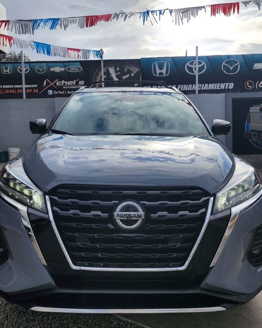 Nissan Kicks gris de frente en el lote de Ixchel's Automotriz, con el letrero del negocio y banderines al fondo